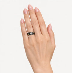 A hand with light pink manicured nails wearing a black ring with a thin gold accent stripe, set against a plain light gray background.