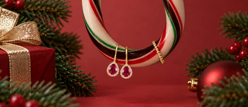 Gold holiday jewelry displayed on a candy cane-shaped stand against a red background, including a heart-shaped pendant necklace, a small gold chain, and a pair of dangling earrings with pink gemstones. Pine branches, red berries, a wrapped gift box, and a red ornament frame the scene.